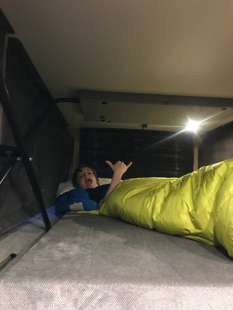 A child makes a shaka sign and shouts in a bright yellow sleeping bag, enjoying a cozy bed setup in an RV rental.