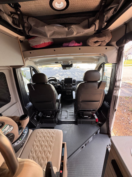 View inside an RV rental looking forward, highlighting the cockpit, two front seats, a child car seat, and overhead storage compartments.