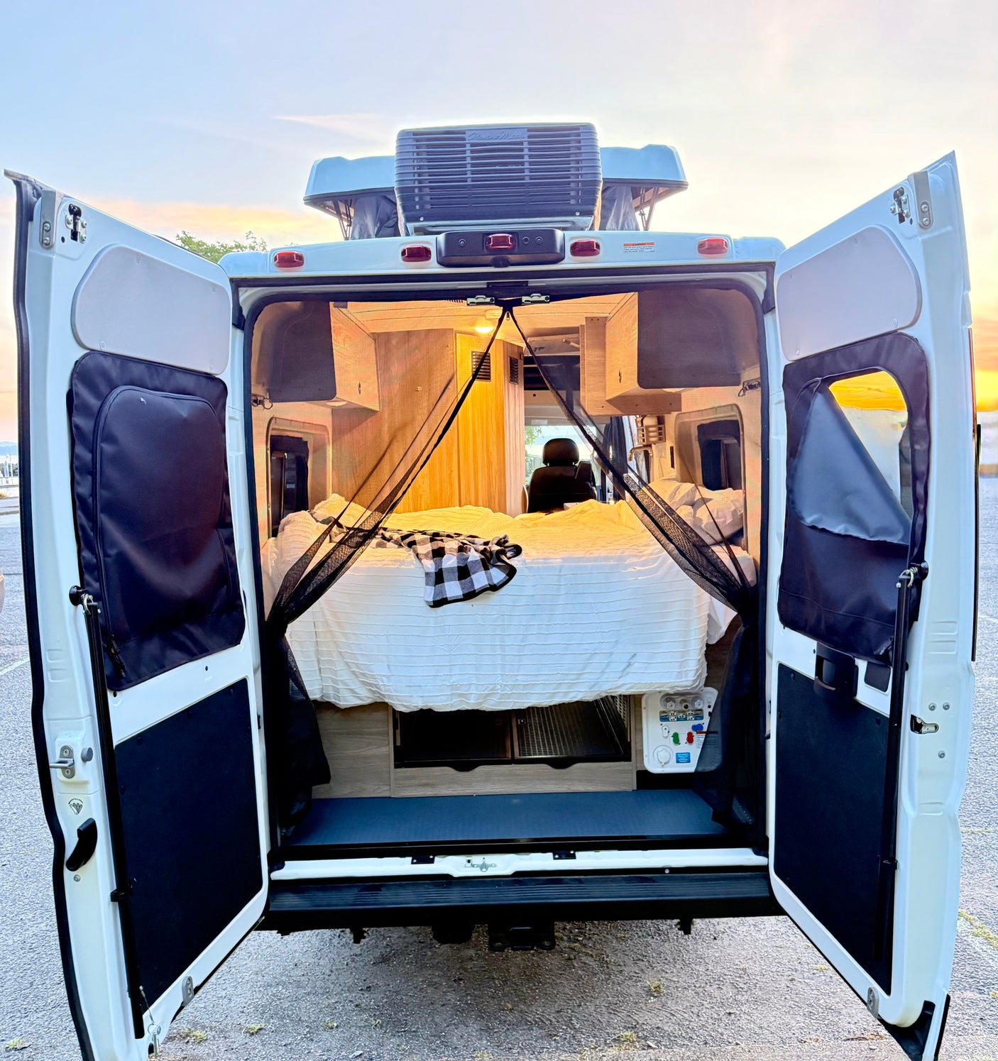 Open rear doors of an RV rental camper van reveal a comfortable bed and mosquito net inside, ready for adventure at sunset.