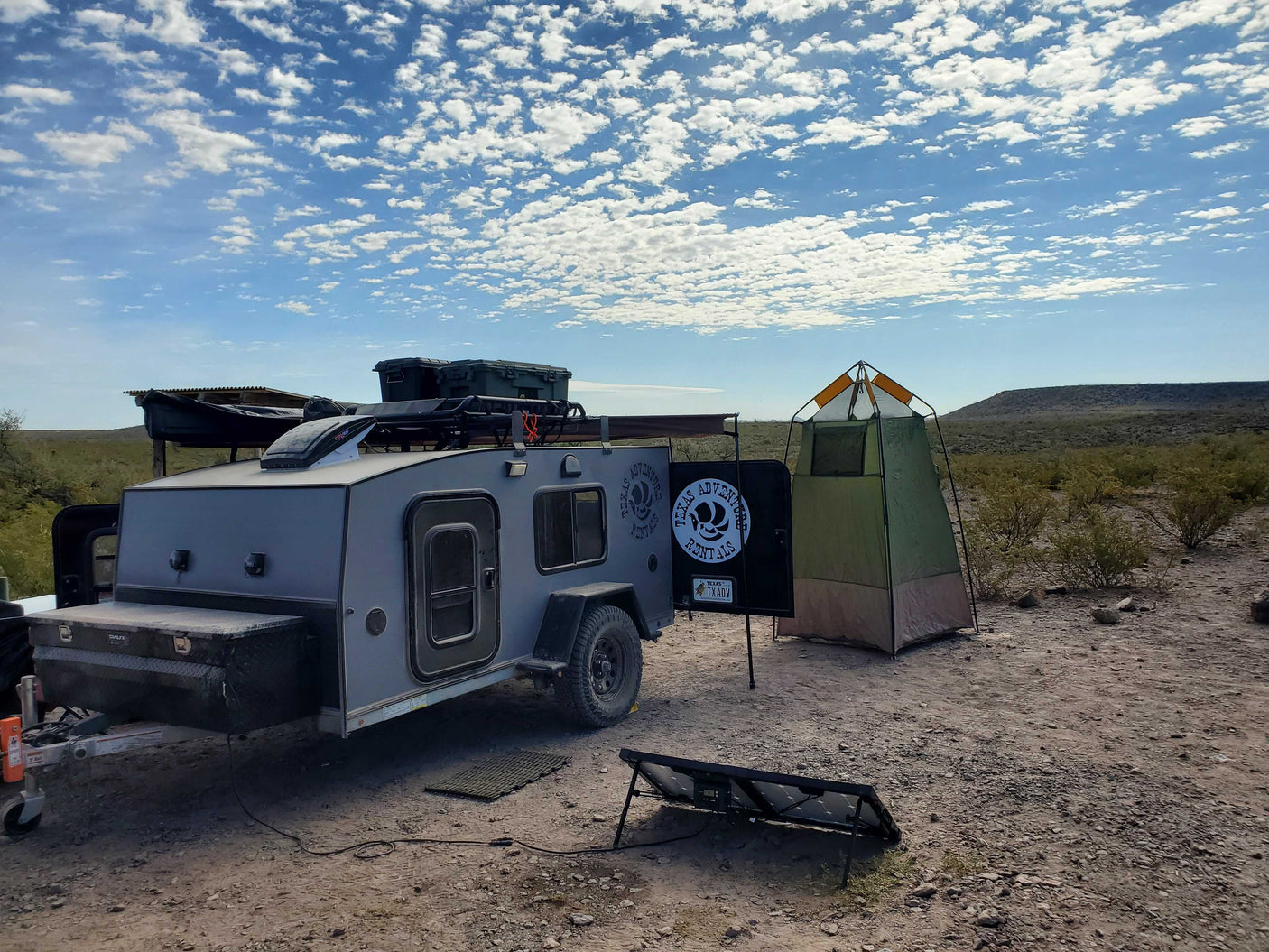 An off-road RV rental, privacy tent, and solar panel are set up for desert camping under a cloudy sky.