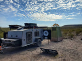 An off-road RV rental, privacy tent, and solar panel are set up for desert camping under a cloudy sky.