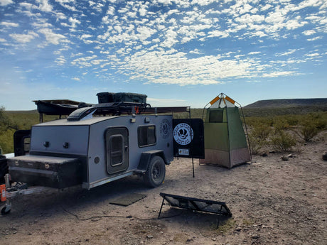 An off-road RV rental, privacy tent, and solar panel are set up for desert camping under a cloudy sky.