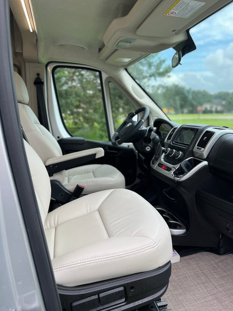 RV rental's front cabin. Cream-colored seats with armrests, steering wheel, and dashboard with infotainment screen.