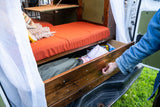 Hand opening a wooden storage drawer under an orange bed cushion in an RV rental.