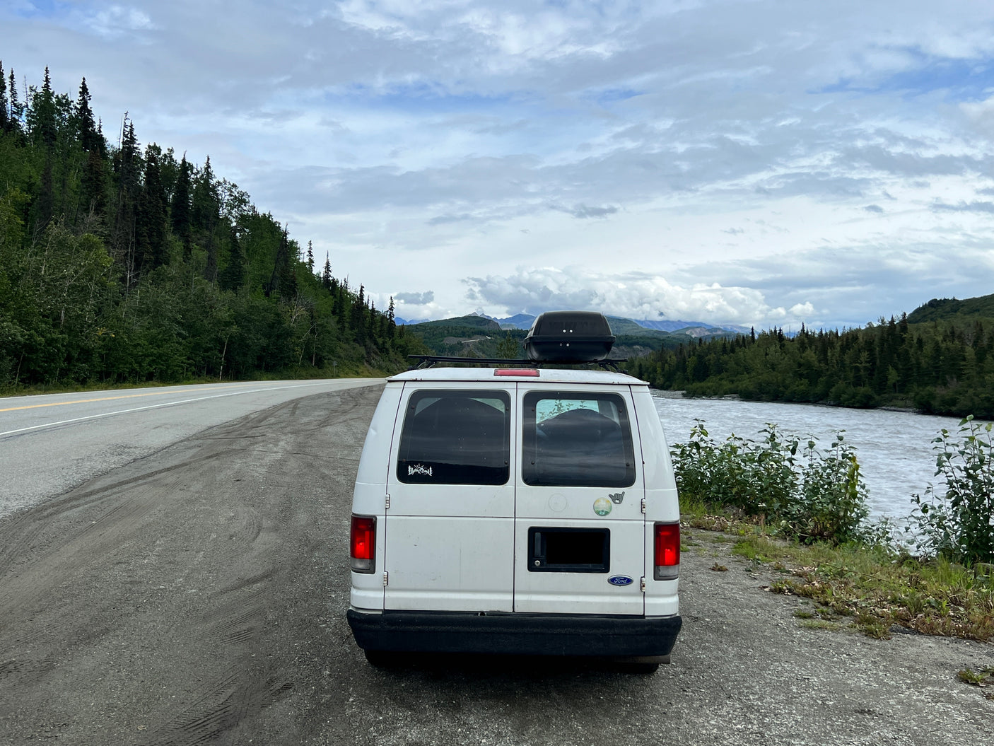 White RV rental van parked beside a river and forest. Scenic road trip stop with mountains and cloudy skies.