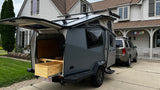 Gray teardrop camper with open rear kitchen/storage, hitched to an SUV in a driveway. Perfect RV rental for adventures!