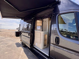 Coastal RV rental: Dark grey camper van parked by a sandy beach with its side door open, revealing a modern interior and extended awning.
