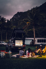 Friends enjoy an outdoor movie projected from a truck, camping under palm trees. An RV rental is visible in the background.