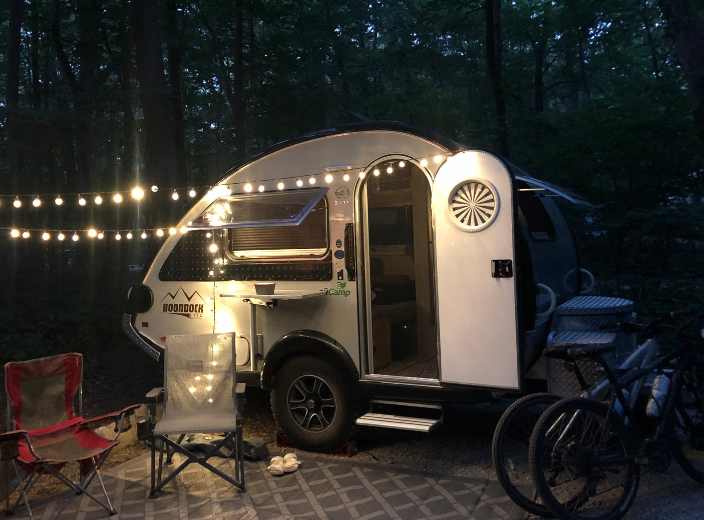 A small 'Boondock Lite' **RV rental** illuminated by string lights at dusk in a wooded campsite, with camping chairs and bikes.