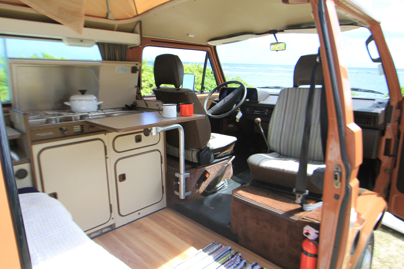 Explore this cozy camper van RV rental interior: kitchen, dining table with mugs, and front seats with stunning ocean views.