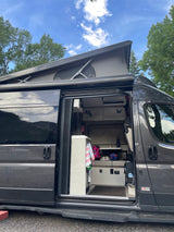 Open side door of an RV rental camper van with pop-top roof, revealing a cozy, decorated interior. Surrounded by trees & blue sky.
