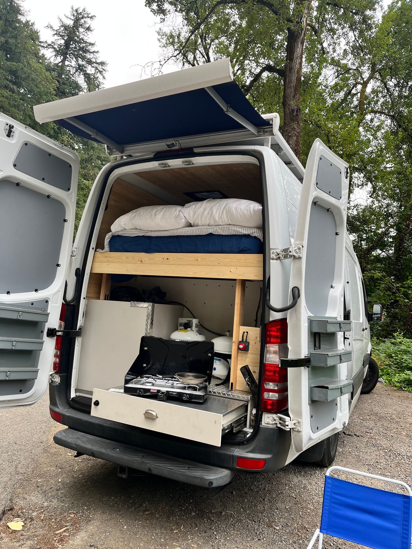 An RV rental camper van, rear open, reveals a compact bed, pull-out kitchen, and extended side awning.