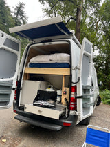 An RV rental camper van, rear open, reveals a compact bed, pull-out kitchen, and extended side awning.
