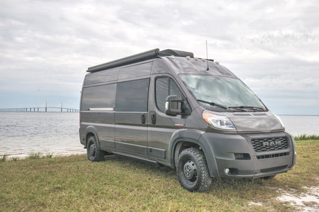 Scenic RV rental: A grey Ram ProMaster van parked by calm waters with a distant bridge under cloudy skies.