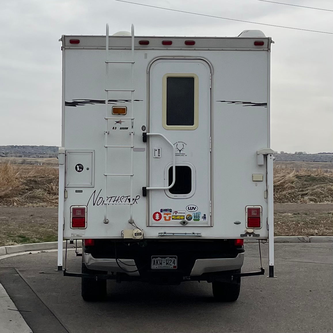 A white truck camper, often an RV rental, stands ready for travel. The rear shows a ladder, a door, and a Colorado license plate.