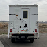 A white truck camper, often an RV rental, stands ready for travel. The rear shows a ladder, a door, and a Colorado license plate.