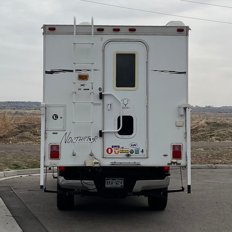 A white truck camper, often an RV rental, stands ready for travel. The rear shows a ladder, a door, and a Colorado license plate.