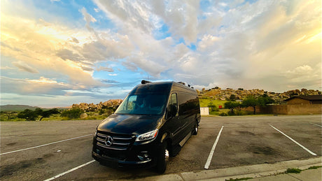 Black Sprinter rv rental van parked in a scenic lot with rocky hills & a dramatic sunset sky. Ready for adventure!