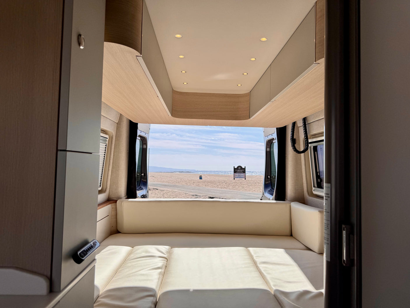 Cozy RV rental interior with light wood and cream cushions, offering a stunning beach and ocean view from the open back doors.