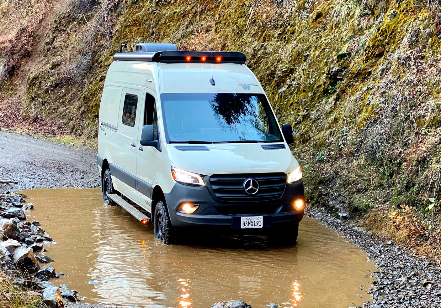 A white Mercedes-Benz Sprinter RV rental drives through a large muddy puddle on a dirt road, ready for off-road adventure.