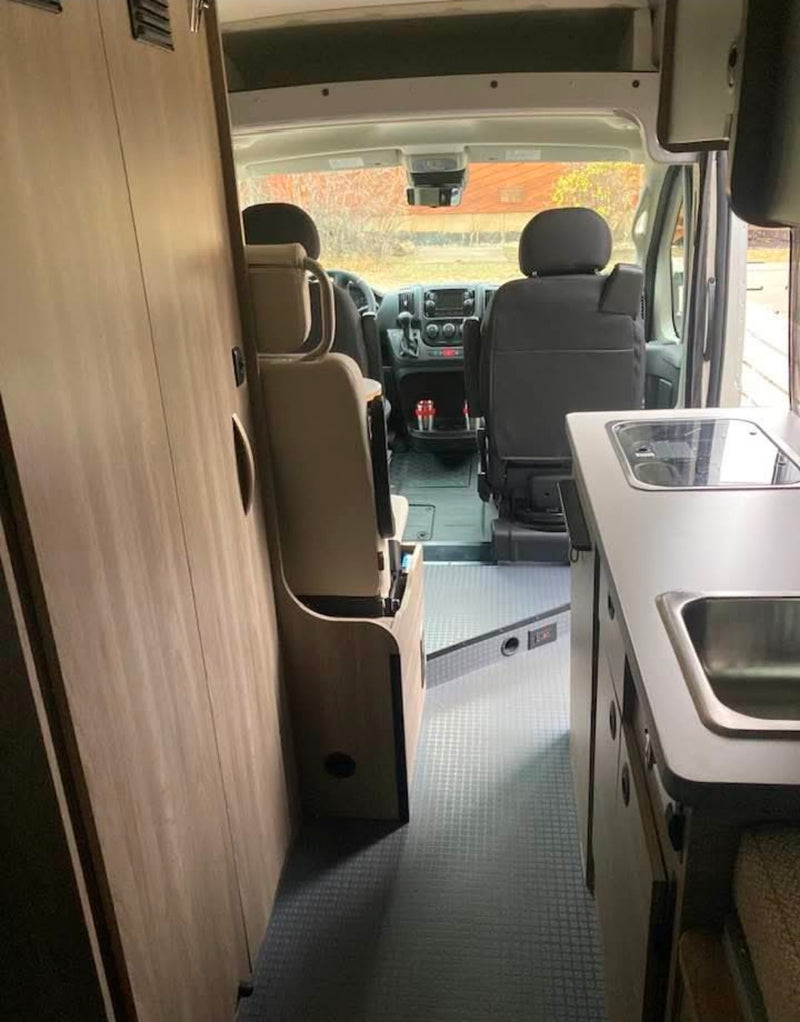  stands next to a red RV rental camper van by a river in a forest.|This RV rental features a well-equipped campervan interior with kitchen — photo 6
