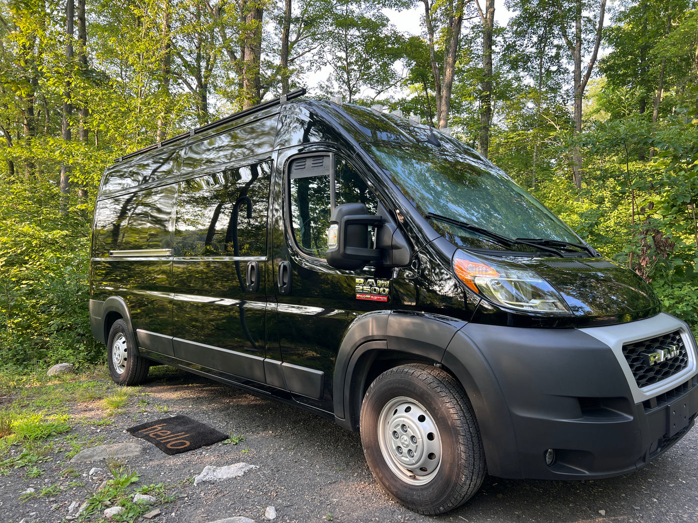 Black Ram ProMaster 2500 RV rental parked in a lush forest, featuring a 'hello' mat. Perfect for outdoor travel.