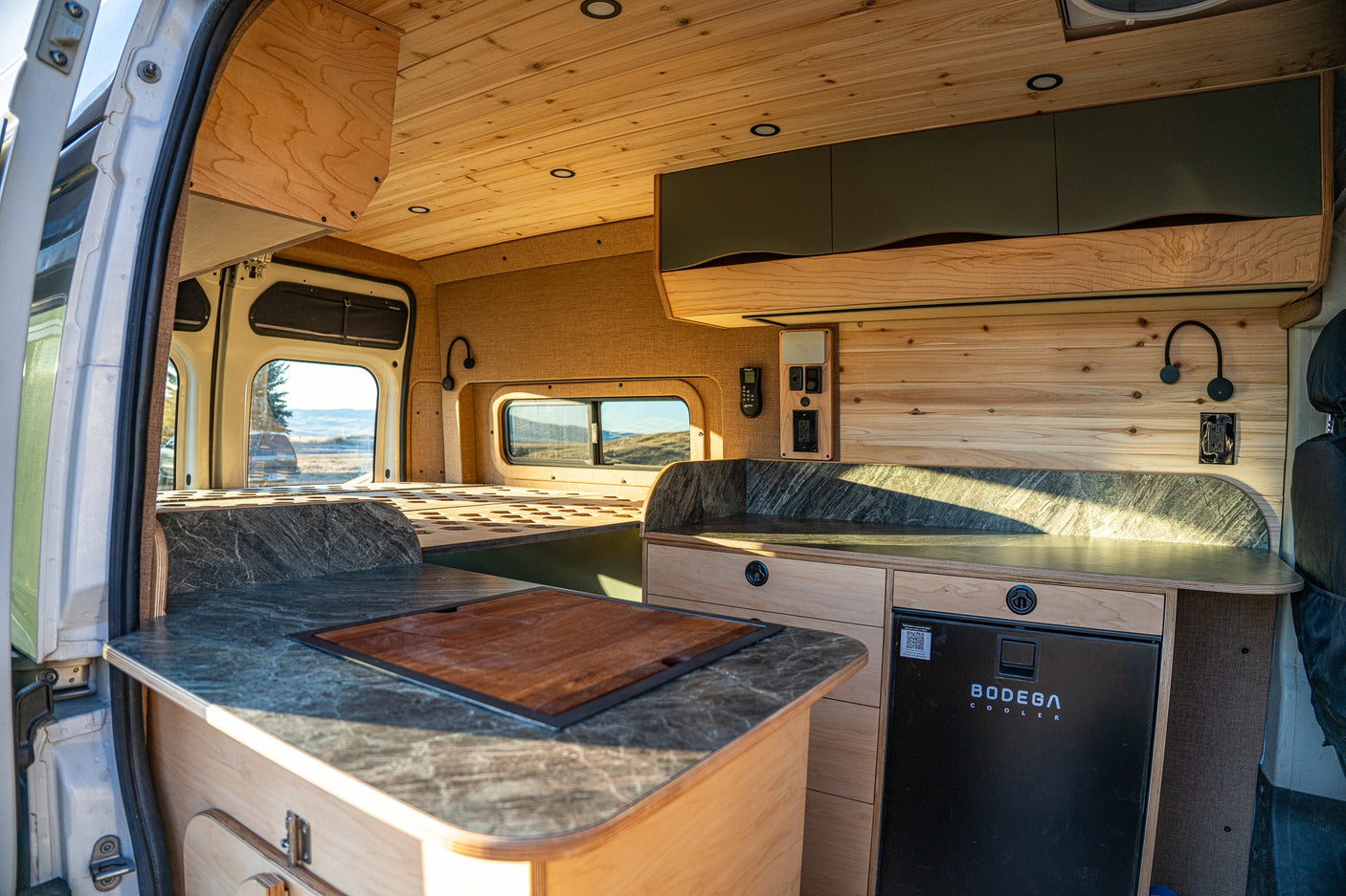 Bright, modern rv rental camper van interior with custom wood, kitchen counter, fridge, and bed area visible for travel.