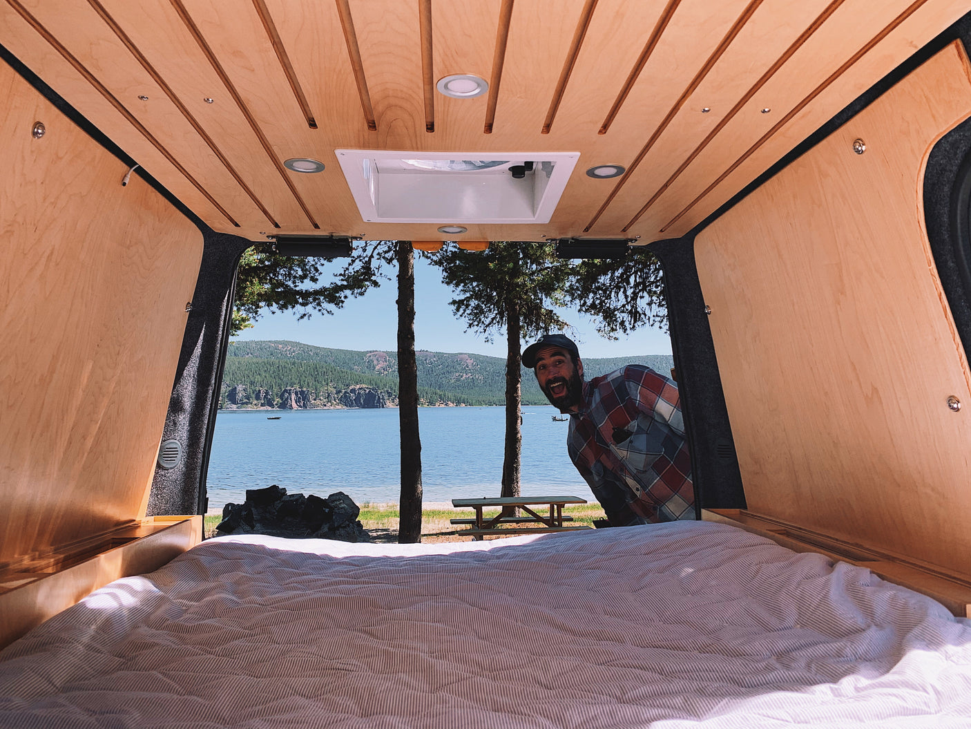 Inside a cozy RV rental, a smiling man looks in from a lakeside campsite with a mountain view.