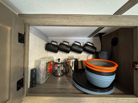 Inside an RV rental cabinet: organized black mugs, coffee makers, and stackable blue/orange dishes, plus a first-aid kit.