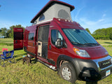Red Winnebago Solis rv rental with pop-top and side door open, a dog, and chairs on grass. Bright outdoor camping scene.