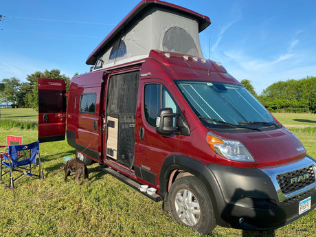 Red Winnebago Solis rv rental with pop-top and side door open, a dog, and chairs on grass. Bright outdoor camping scene.