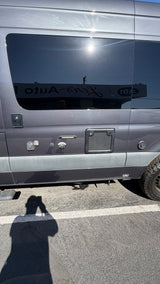 Side of a gray camper van for rv rental, showing utility connections, tinted windows reflecting a building, and a person's shadow.