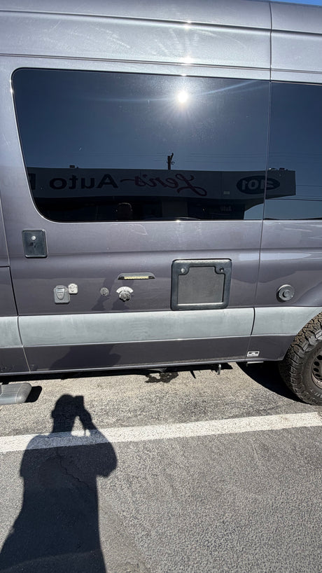 Side of a gray camper van for rv rental, showing utility connections, tinted windows reflecting a building, and a person's shadow.