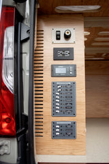 Inside an RV rental: detailed view of a camper van's sophisticated electrical control and power distribution system on wood paneling.
