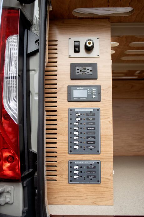 Inside an RV rental: detailed view of a camper van's sophisticated electrical control and power distribution system on wood paneling.