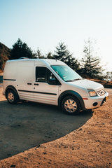 White camper van, ideal for RV rental, parked on a dirt shoulder by a road with scenic trees and mountains.