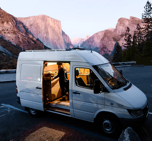 An illuminated white camper van, perfect for **rv rental**, with a person inside at dusk, set against grand mountains.