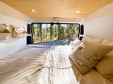 Serene RV rental bedroom, featuring a cozy bed & hanging storage, with a stunning forest view from the rear.