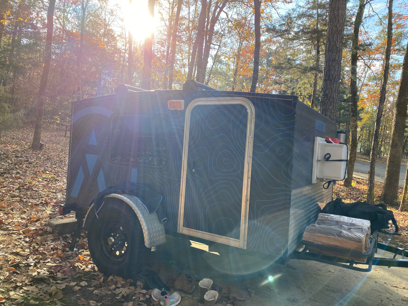 Teardrop RV rental with topographic design, parked in a sunny, wooded campsite with fallen leaves & firewood.