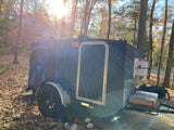 Teardrop RV rental with topographic design, parked in a sunny, wooded campsite with fallen leaves & firewood.