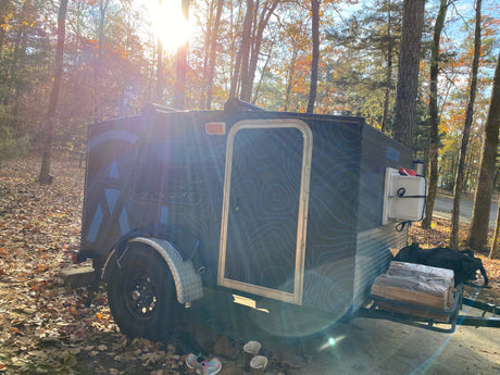 Teardrop RV rental with topographic design, parked in a sunny, wooded campsite with fallen leaves & firewood.