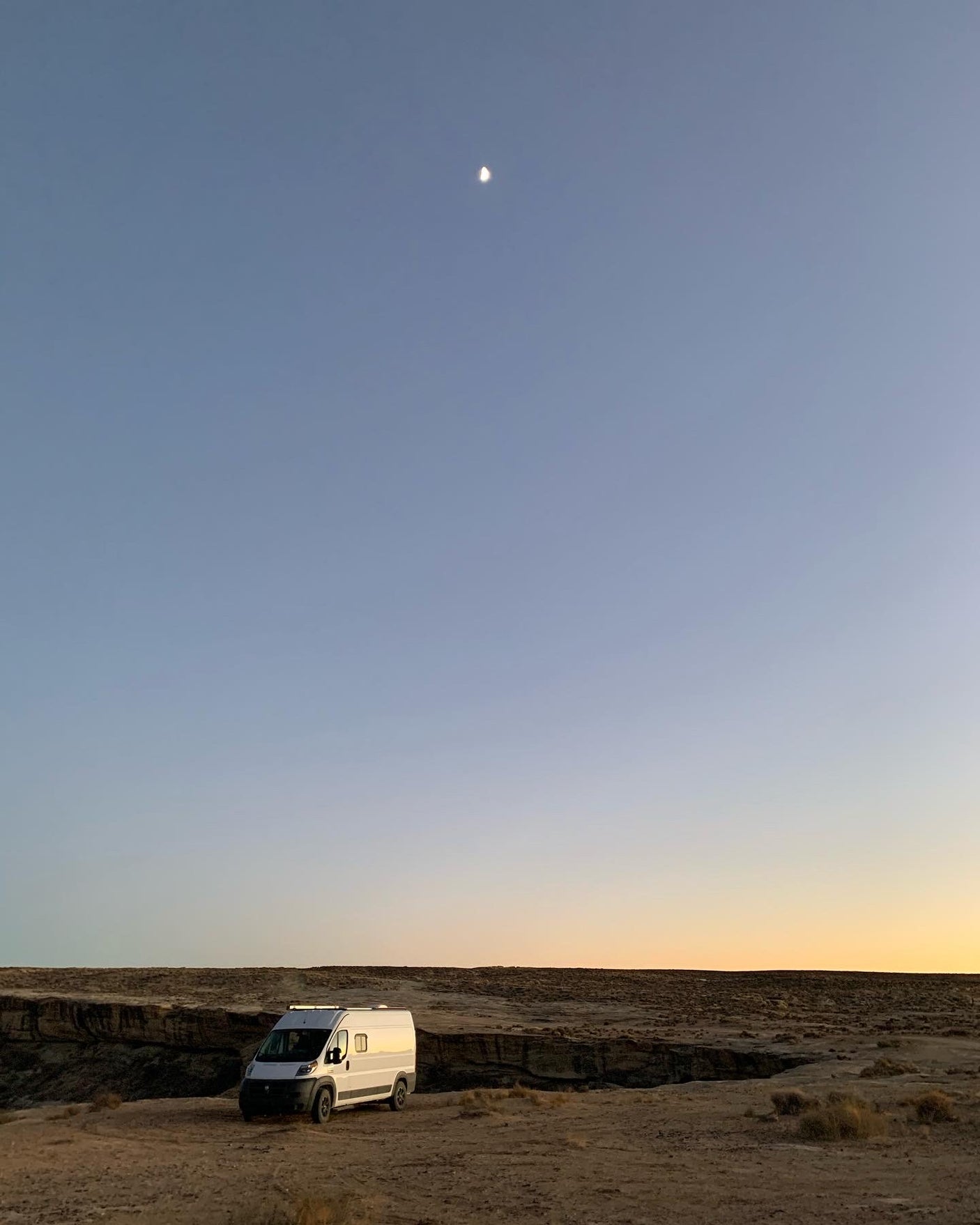A white camper van rests in a vast desert under a clear sky with a crescent moon & sunset colors. Explore nature with an RV rental.