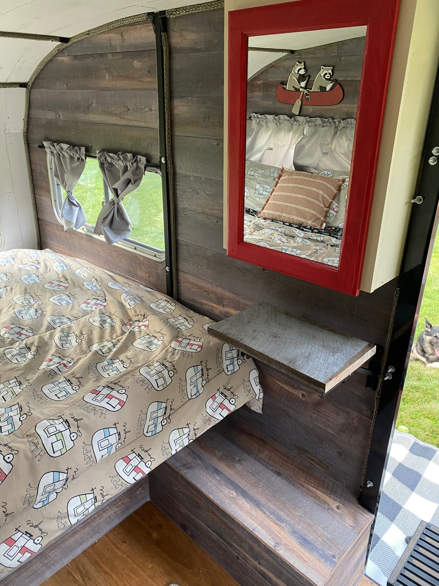 Explore this comfy RV rental! Features "Happy Camper" bedding, wood interior, a unique mirror, and a dog visible outside.