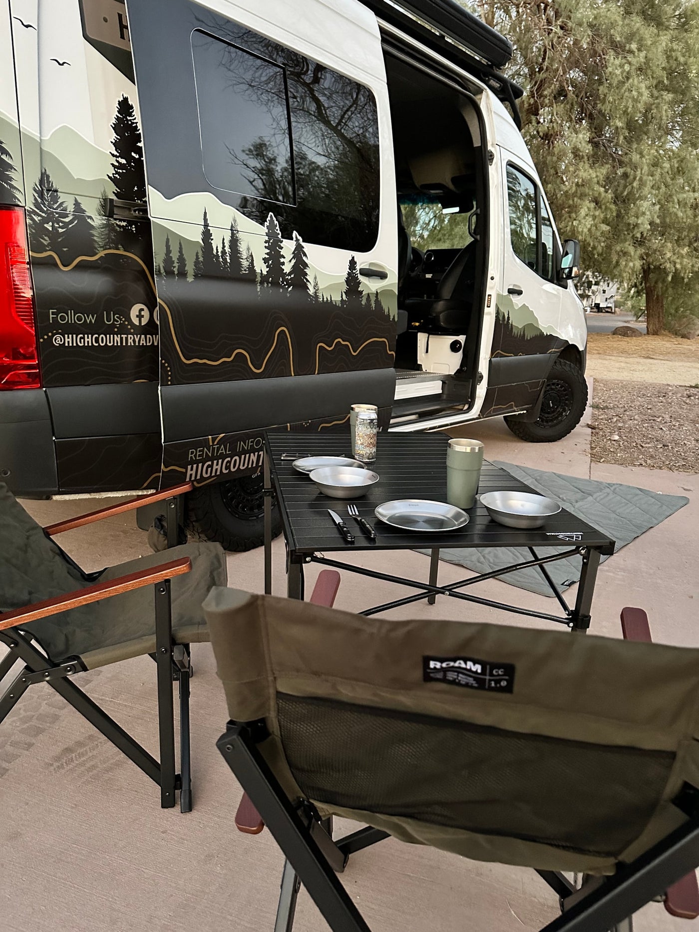 This RV rental van, with custom mountain graphics, offers an outdoor camping setup: chairs, table, and kitchenware. — photo 2