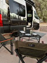 This RV rental van, with custom mountain graphics, offers an outdoor camping setup: chairs, table, and kitchenware.