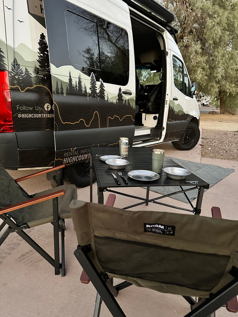 This RV rental van, with custom mountain graphics, offers an outdoor camping setup: chairs, table, and kitchenware. — photo 2