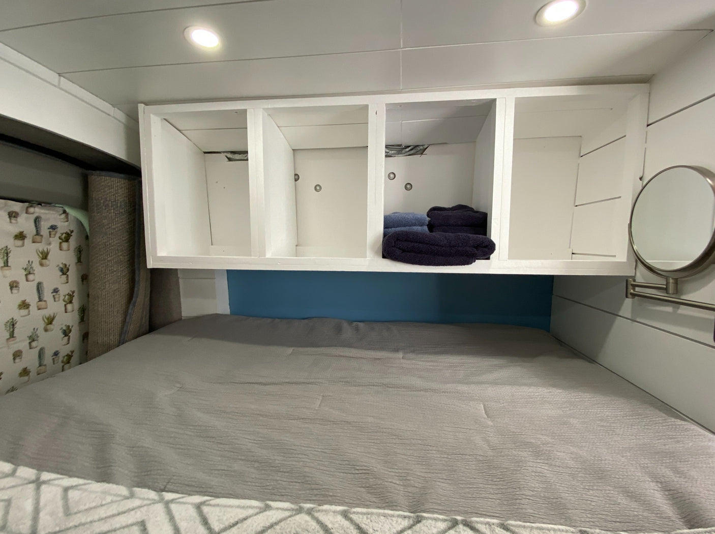 Inside an RV rental: a cozy bed, white storage cubbies with folded towels, and a blue accent wall. Compact and functional space.