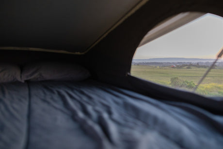 Inside an RV rental: a comfy bed awaits, offering a scenic view of green fields, a red barn, and hazy mountains.
