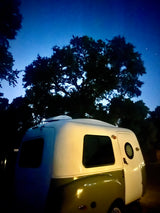 A small, retro rv rental parked at night under dark trees and a starry blue sky, ready for adventure.