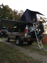 A person climbs a ladder to a rooftop tent on a Land Rover Discovery.